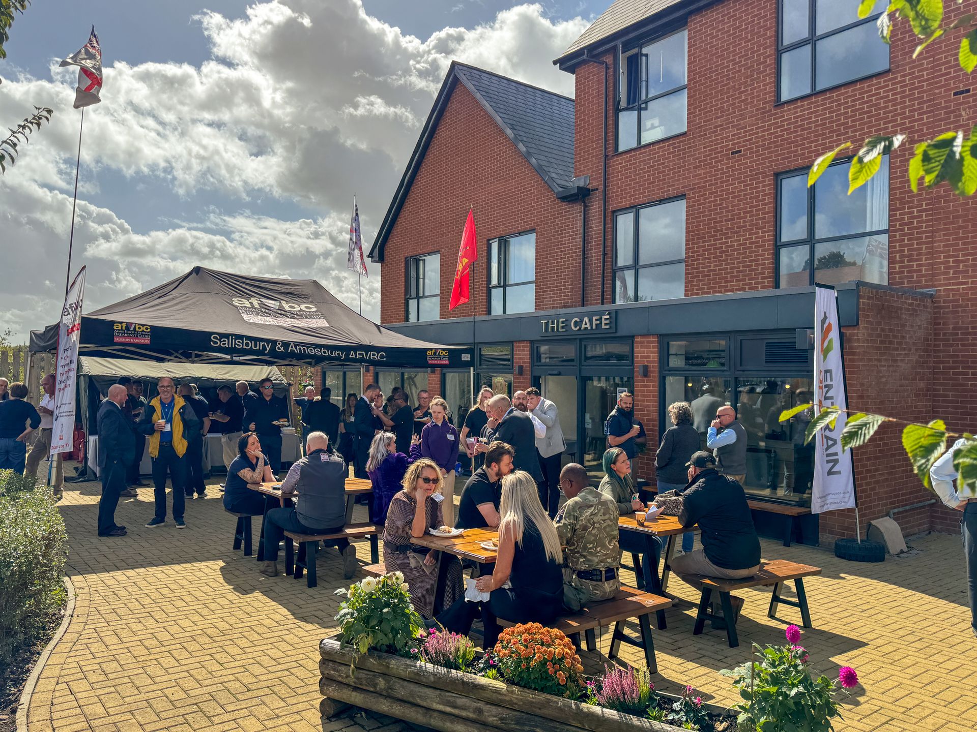 Outdoor community event at The Café, Erskine House with people at tables and military flags